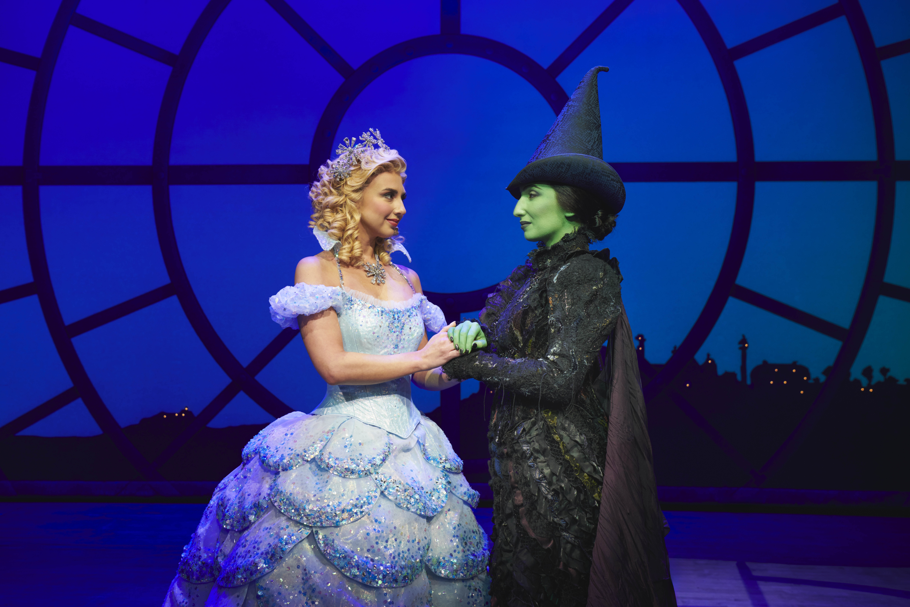 Glinda and Elphaba holding hands looking at one another. Glinda wears a pale blue full skirted ballgown and tiara, Elphaba wears a black ruffled dress, black cape and black witches hat, she has green skin.